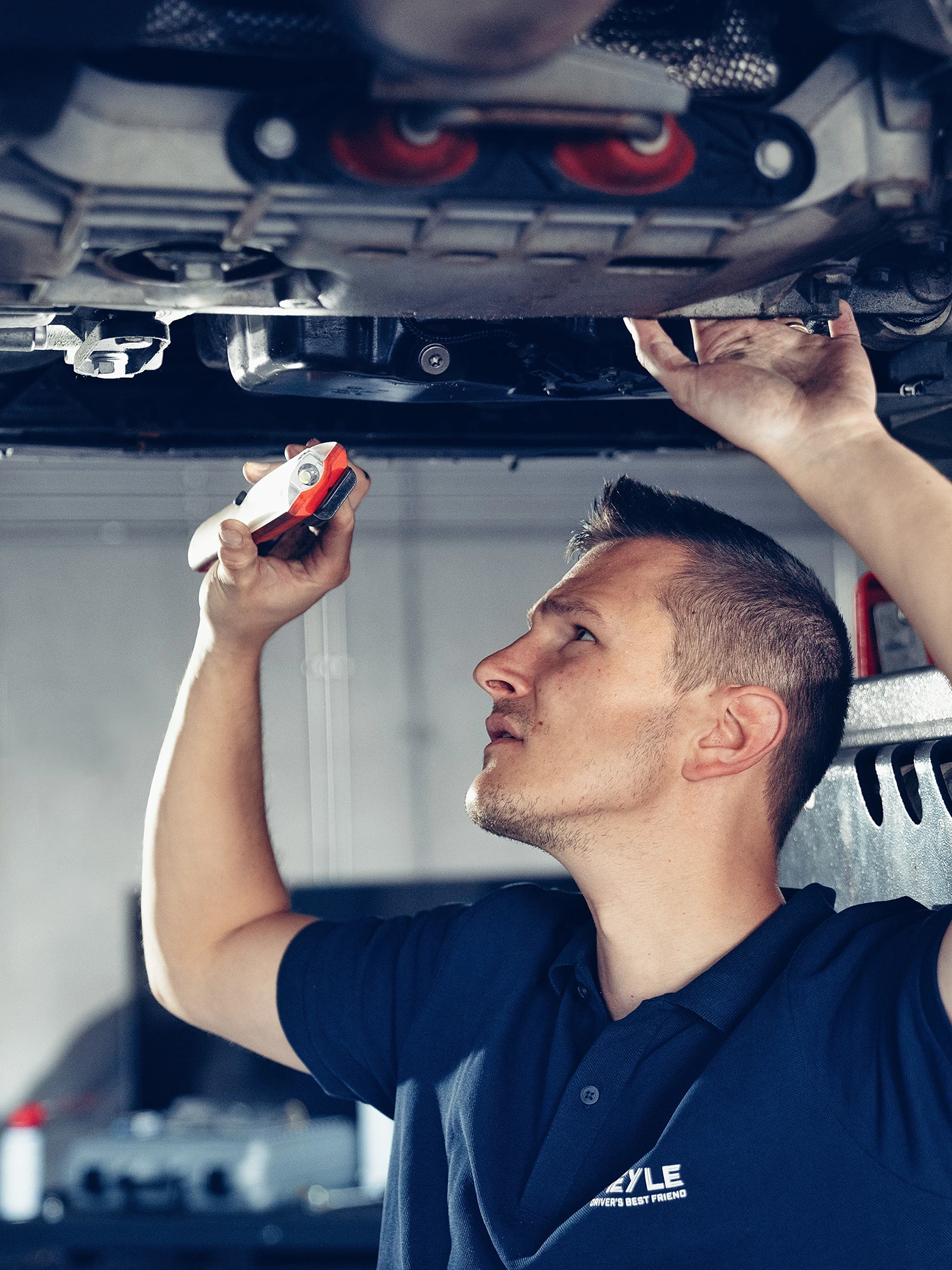 Mechaniker steht unter einem Auto was auf der Hebebühne steht und guckt sich ein Antriebsteil ganz genau an.