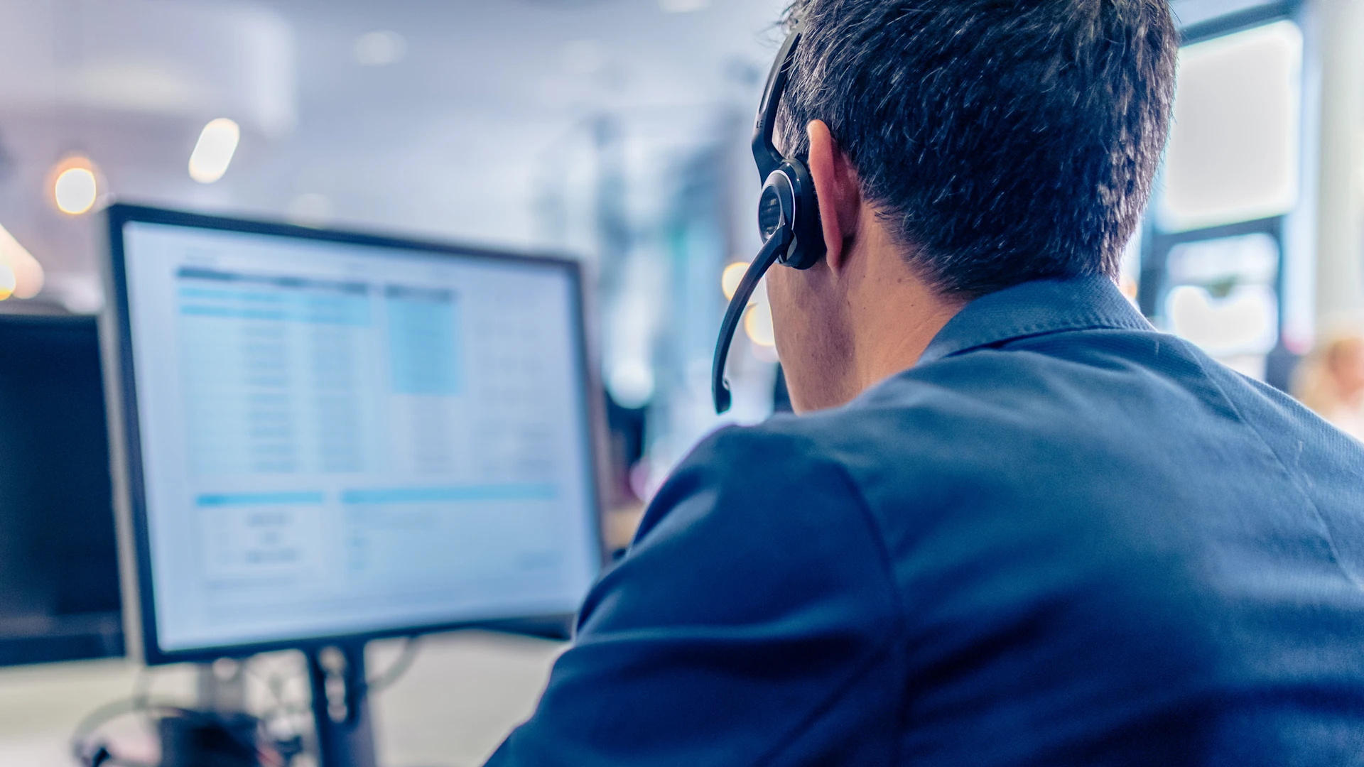Person from behind with headset, head in focus, in the background is a monitor in the blur.