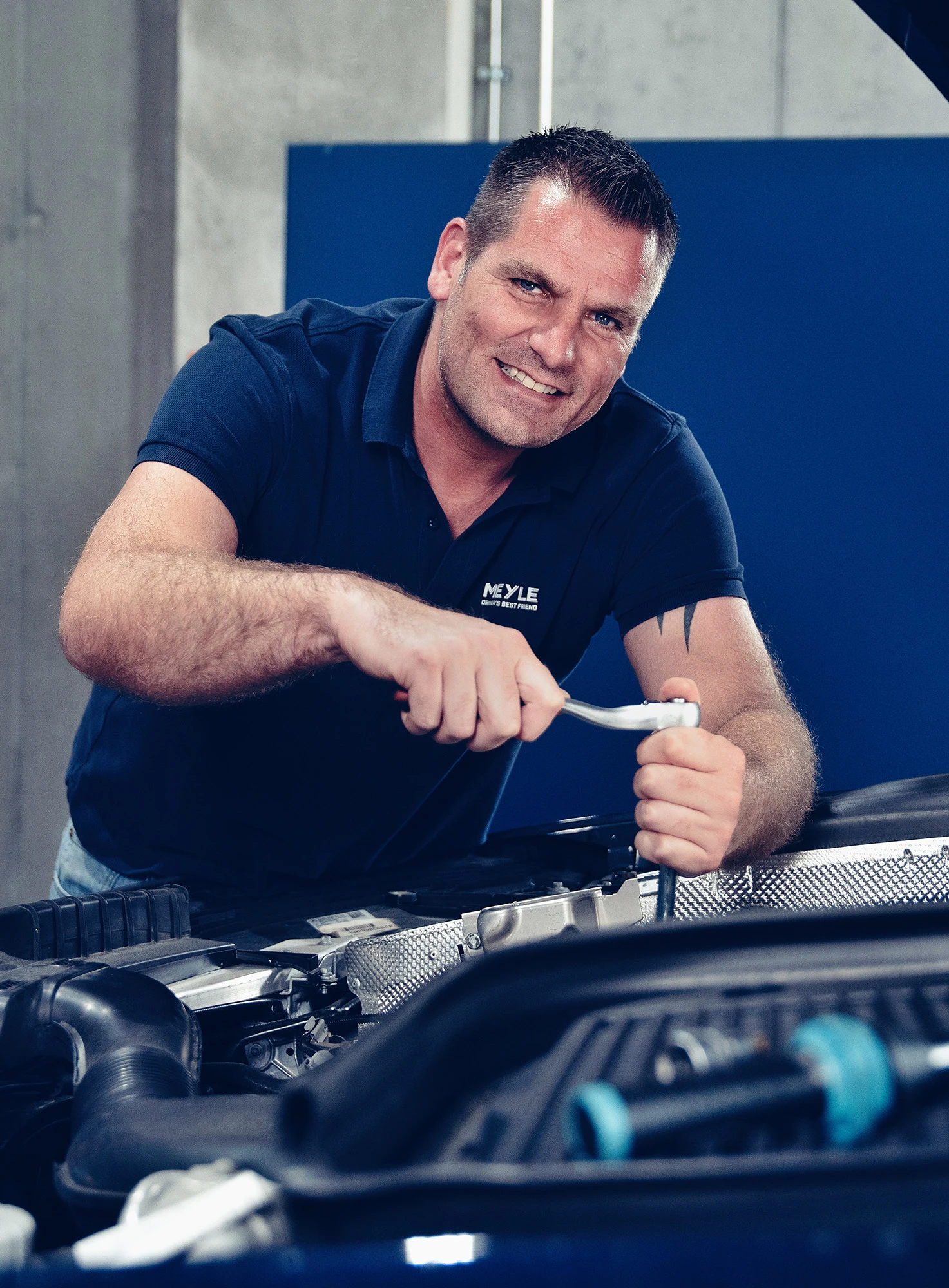 Mechanic working in the engine compartment and smiling at the camera.